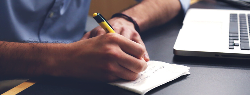 Person writing on a tissue paper with a black pen. A MacBook is angled towards him.