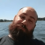 A photo of Chris Huffstutter looking at the camera and smiling while tilting his head. A water body is visible in the background.