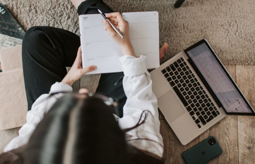 A top-down photo of a woman with black hair, sitting next to an Apple MacBook and an iPhone, while holding a pen and a planner.