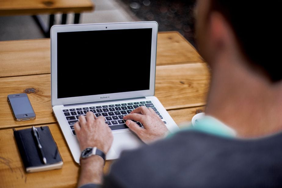 Photo of a person typing something on his Apple computer. A phone and a planner rest next to the laptop.