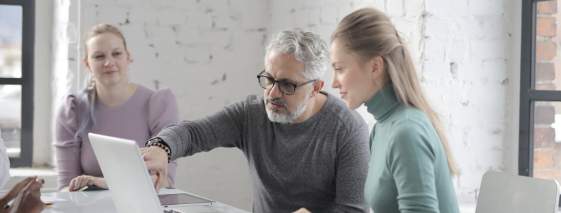 A virtual assistant team working and training together. One person overlooks as another is being taught something on the laptop.