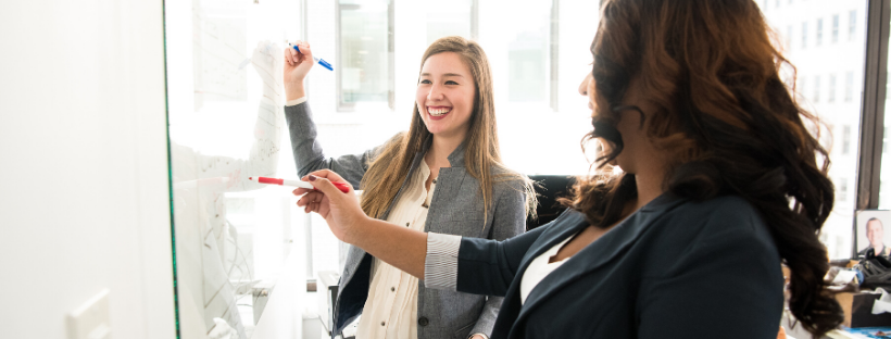 Two women discussing something joyfully while working together on a whiteboard with red and blue markers.