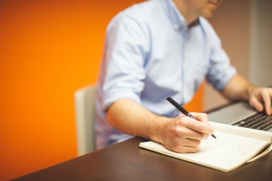A decorative image of an office worker holding a pen to his notebook while working on his laptop with the other.