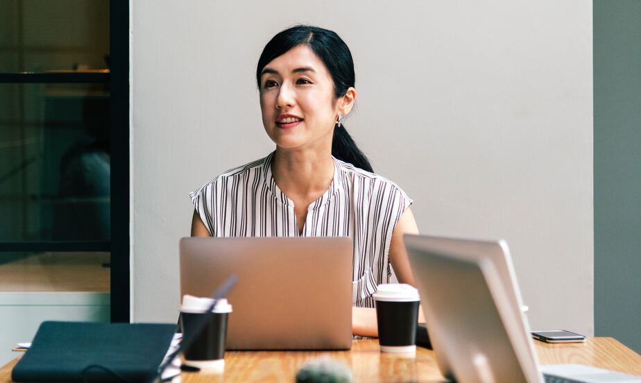An illustrative photo of a woman looking sideways, while sitting at her desk with a laptop among other people.